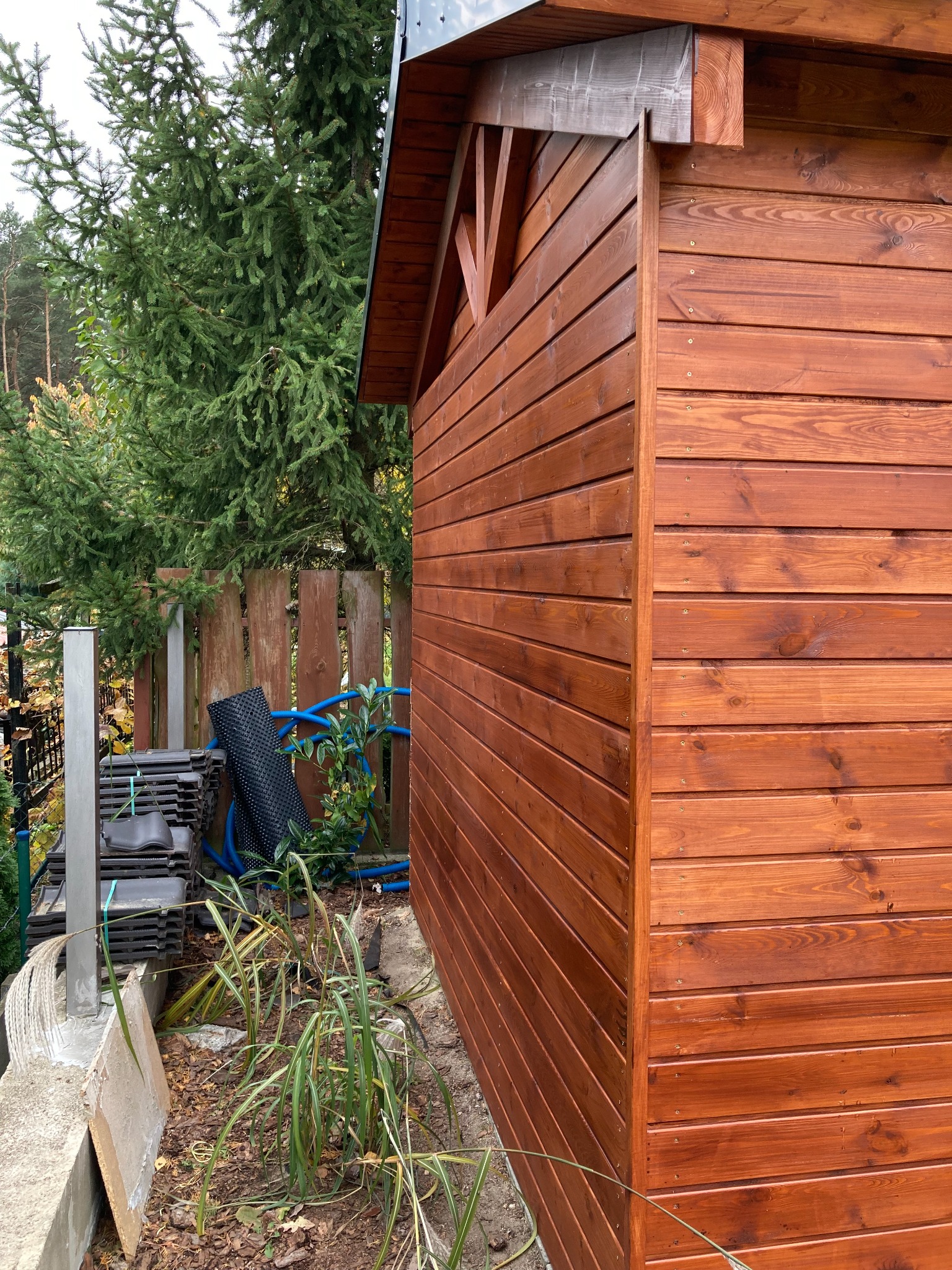 Ściana zewnętrzna nowej, drewnianej konstrukcji z widocznym usłojeniem i detalami połączeń, obok leżą dachówki i rura nawadniająca, w tle drzewa iglaste.