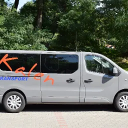 Szary van z pomarańczowym napisem 'Kaleh' i niebieskim 'TRANSPORT' na boku, zaparkowany na chodniku z kostki brukowej, z zielonymi drzewami w tle.