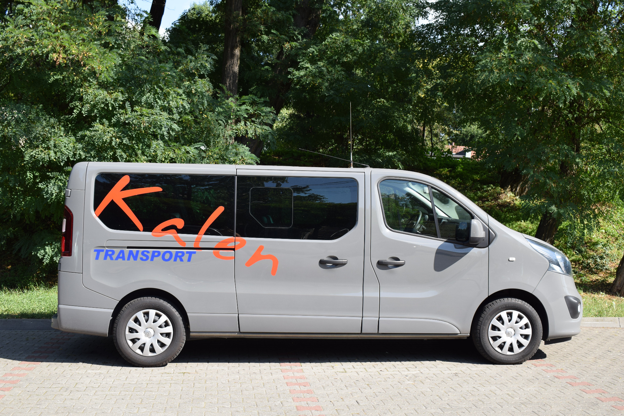 Szary van z pomarańczowym napisem 'Kaleh' i niebieskim 'TRANSPORT' na boku, zaparkowany na chodniku z kostki brukowej, z zielonymi drzewami w tle.