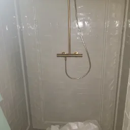 Płytki ścienne w kabinie prysznicowej z zamontowaną złotą armaturą, podłoga zakryta folią ochronną.