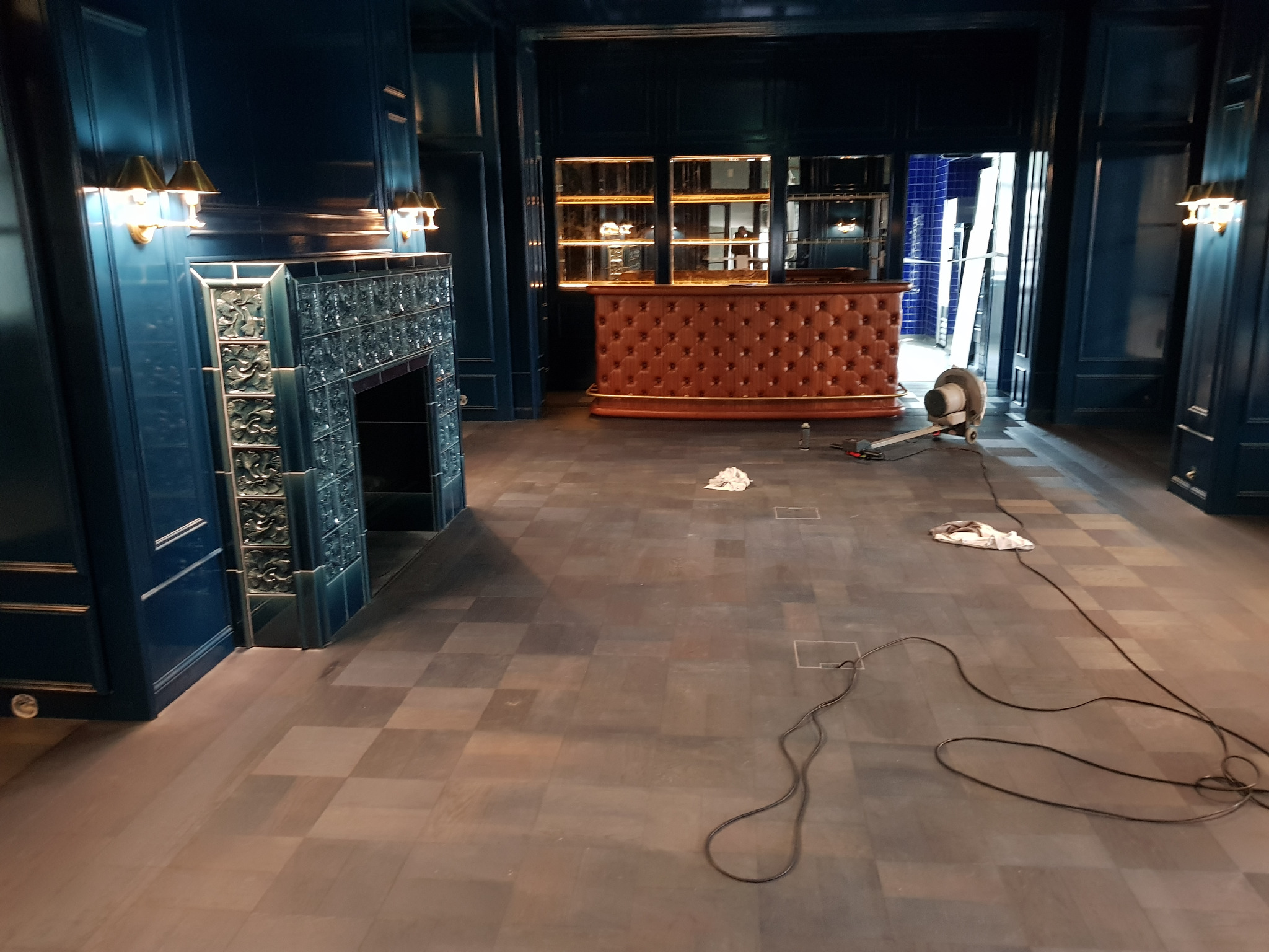 Niebieskie wnętrze z płytkami kominkowymi, drewnianą podłogą w trakcie czyszczenia maszyną, oraz barem w tle.