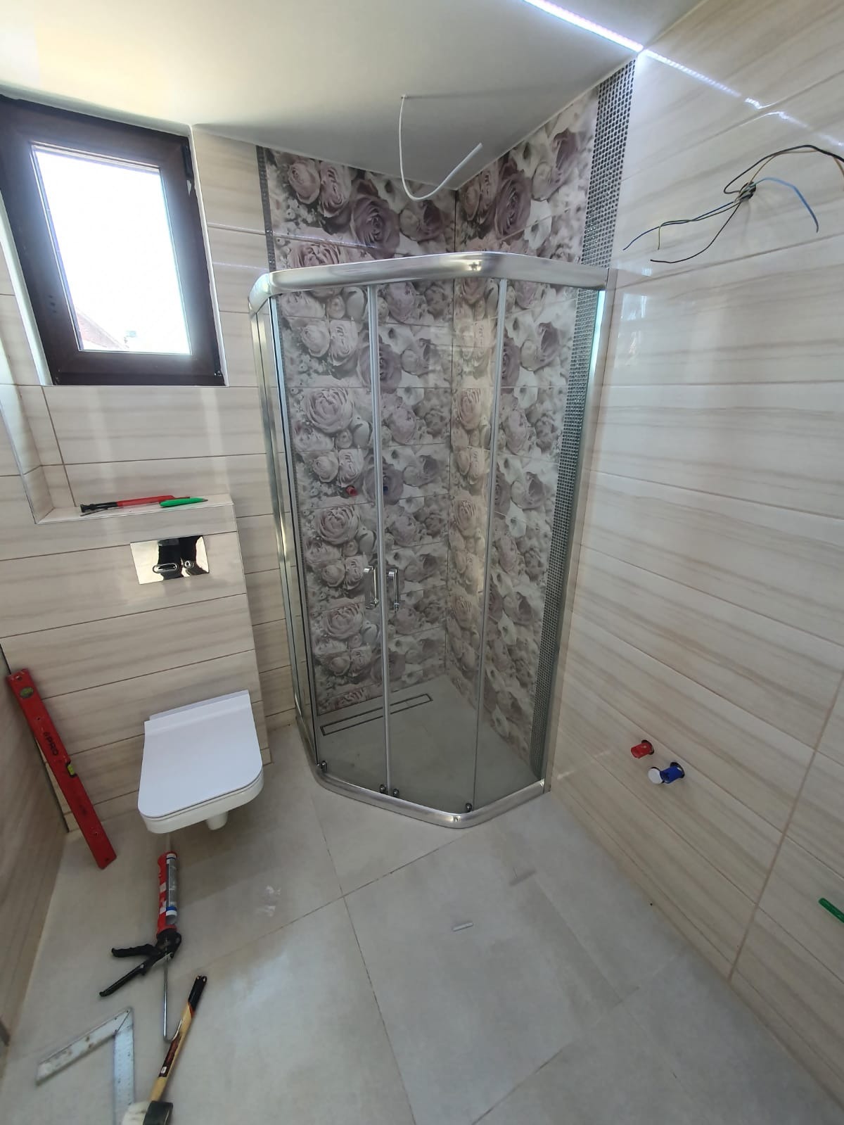 Wykończona łazienka z kabiną prysznicową narożną z motywem kwiatowym na płytkach, wiszącą toaletą i narzędziami na podłodze, poziomica przy ścianie, widoczne kable elektryczne.