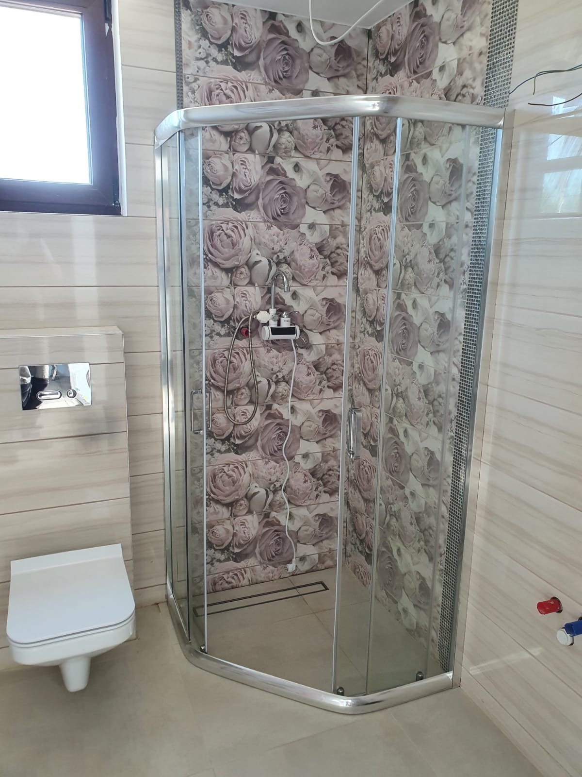 Nowoczesna kabina prysznicowa narożna z chromowanymi profilami i szklanymi ścianami na tle płytek z motywem róż, widoczna toaleta podwieszana i okno.