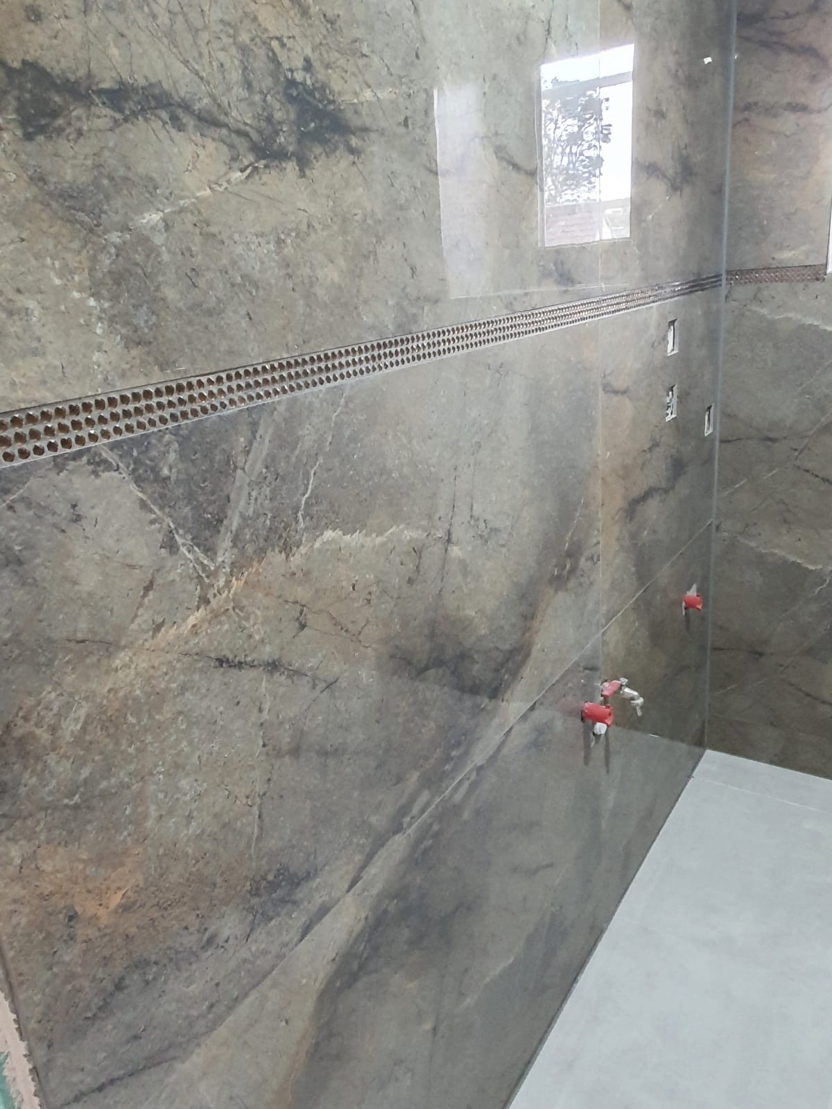 Ułożona ściana z płytek imitujących marmur w odcieniach szarości i brązu, z dekoracyjnym pasem mozaiki i widocznymi plastikowymi krzyżykami do poziomowania płytek.