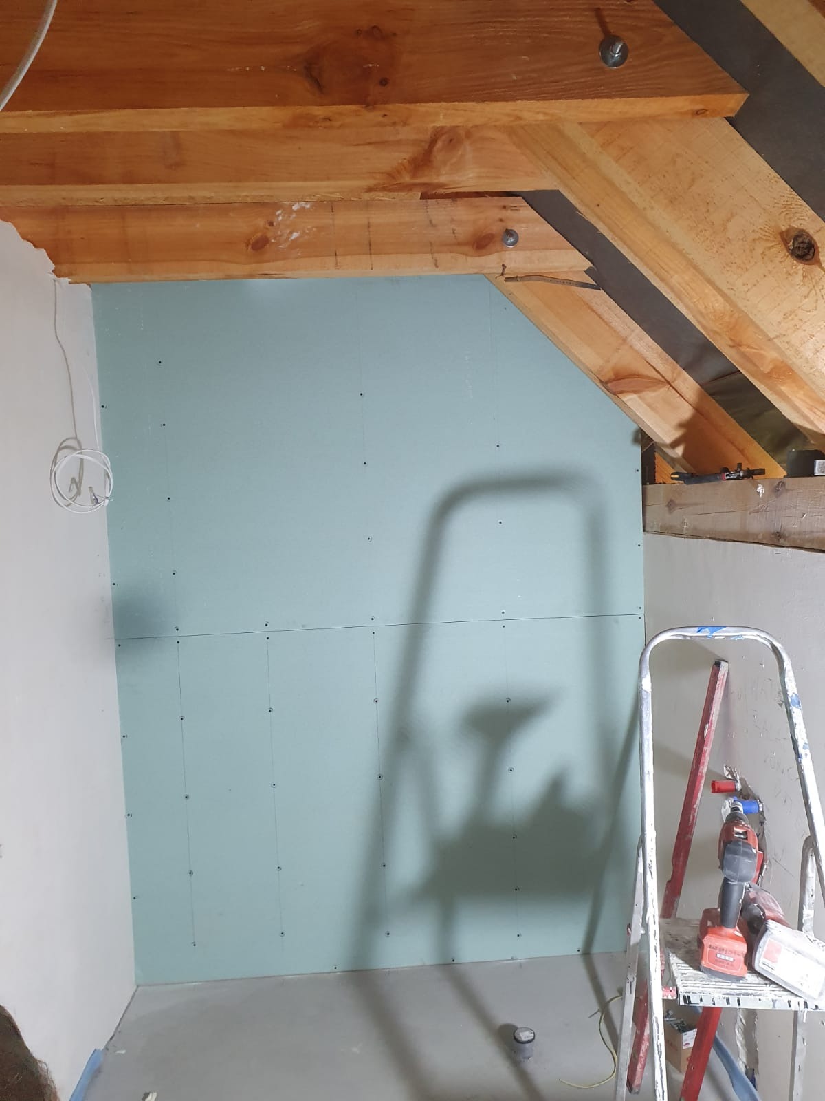 Ściana z zamontowaną płytą gipsowo-kartonową, widoczne drewniane elementy konstrukcyjne poddasza i drabina z narzędziami w tle.