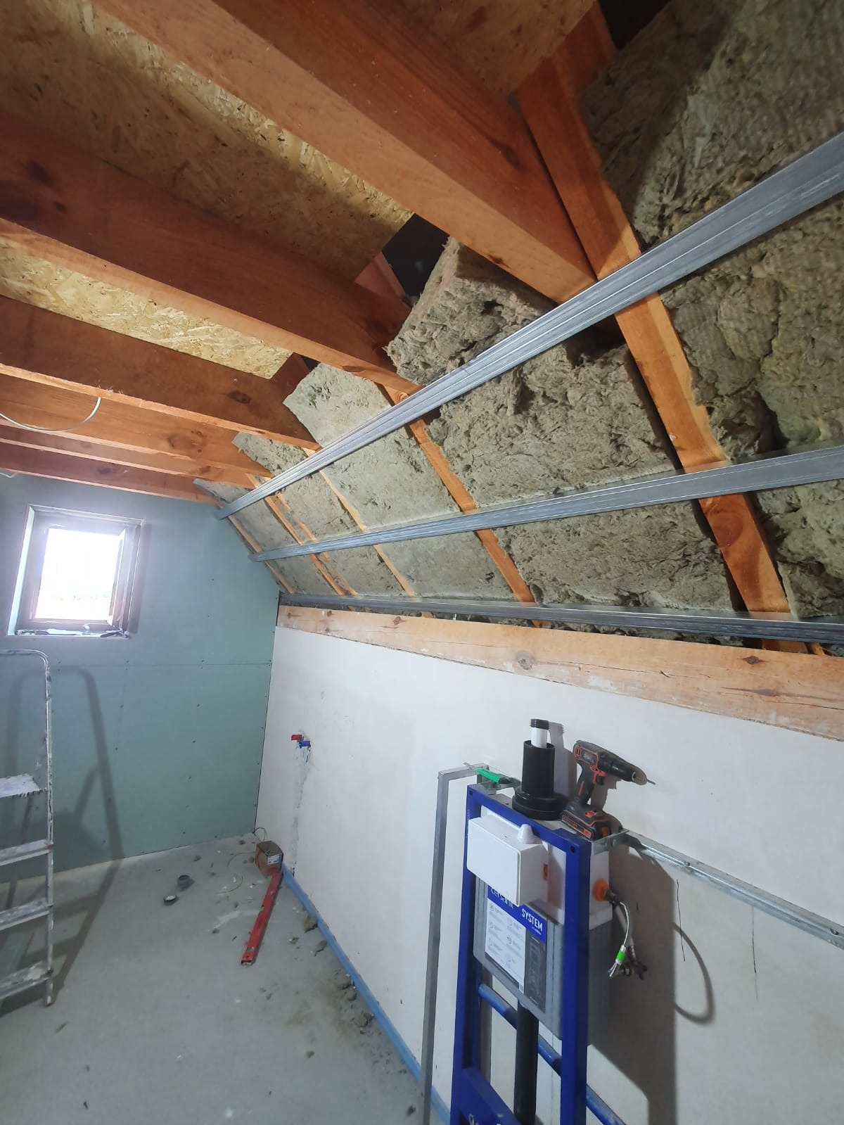 Poddasze w trakcie adaptacji: widoczne drewniane krokwie, izolacja z wełny mineralnej, metalowe profile pod płyty gipsowo-kartonowe oraz stelaż podtynkowy WC z narzędziami.
