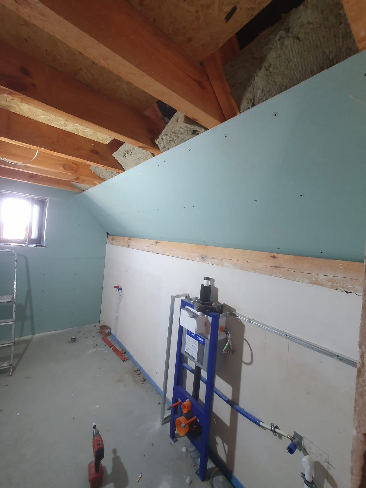 Wnętrze poddasza w trakcie wykańczania, widoczne płyty kartonowo-gipsowe na skośnej ścianie, izolacja z wełny mineralnej pod drewnianą konstrukcją dachu, stelaż podtynkowy WC oraz narzędzia...