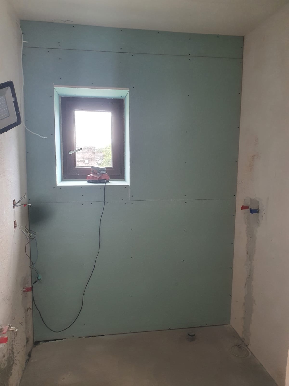 Montaż płyt kartonowo-gipsowych w pomieszczeniu z oknem, widoczne przewody elektryczne i narzędzie na parapecie, surowy beton na podłodze.