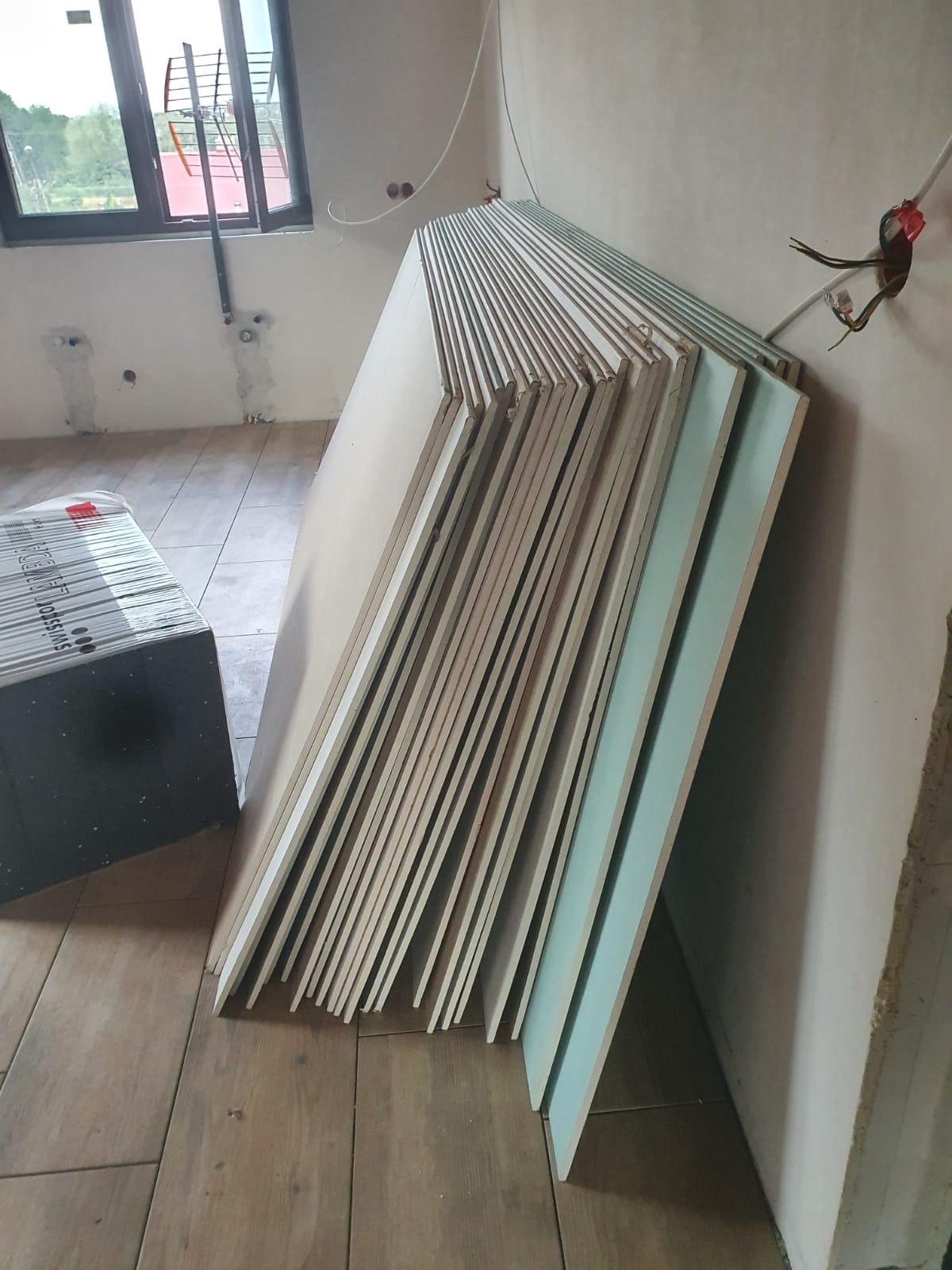 Stos płyt kartonowo-gipsowych, w tym płyty ognioodporne (zielone), opartych o ścianę w surowym wnętrzu z drewnopodobną podłogą i widocznymi instalacjami elektrycznymi.
