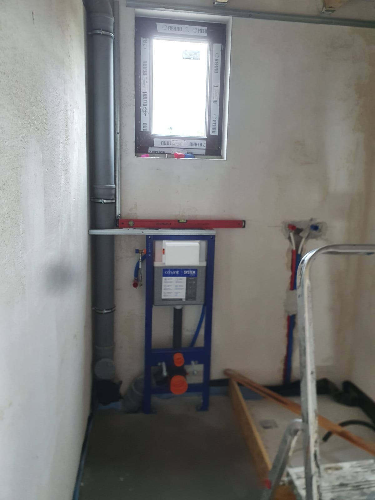 Stelaż podtynkowy WC marki Cersanit zamontowany w surowym wnętrzu łazienki, widoczne rury instalacyjne, okno z naklejkami producenta i poziomica. Ściany przygotowane do dalszych prac wykończeniowych.