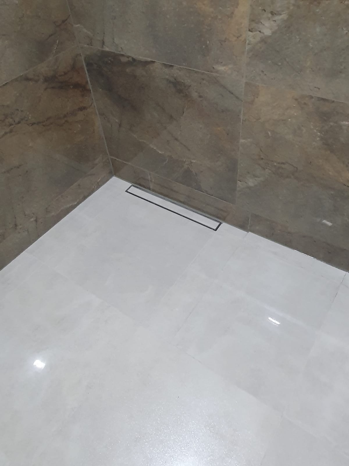 Wykończony narożnik łazienki z szarymi płytkami imitującymi kamień na ścianach i jasnymi płytkami na podłodze, z liniowym odpływem podłogowym.