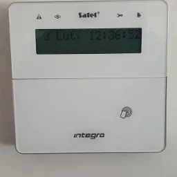 Biały panel sterowania alarmu Satel Integra z wyświetlaczem LCD pokazującym datę 3 lutego i godzinę 12:36:52 oraz ikonami ostrzeżeń i klucza.
