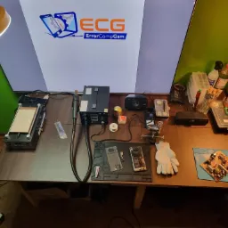 Stanowisko pracy z rozłożonym telefonem, płytką drukowaną, narzędziami i sprzętem serwisowym, oświetlone lampą, z logo firmy ErrorCompGsm na monitorze w tle.