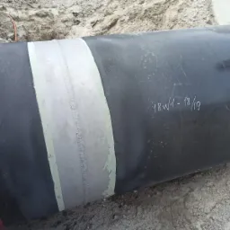 Fragment rury po piaskowaniu i malowaniu, z widocznymi spawami zabezpieczonymi szpachlą i oznaczeniami '18W1-18/19', leżącej na piasku.