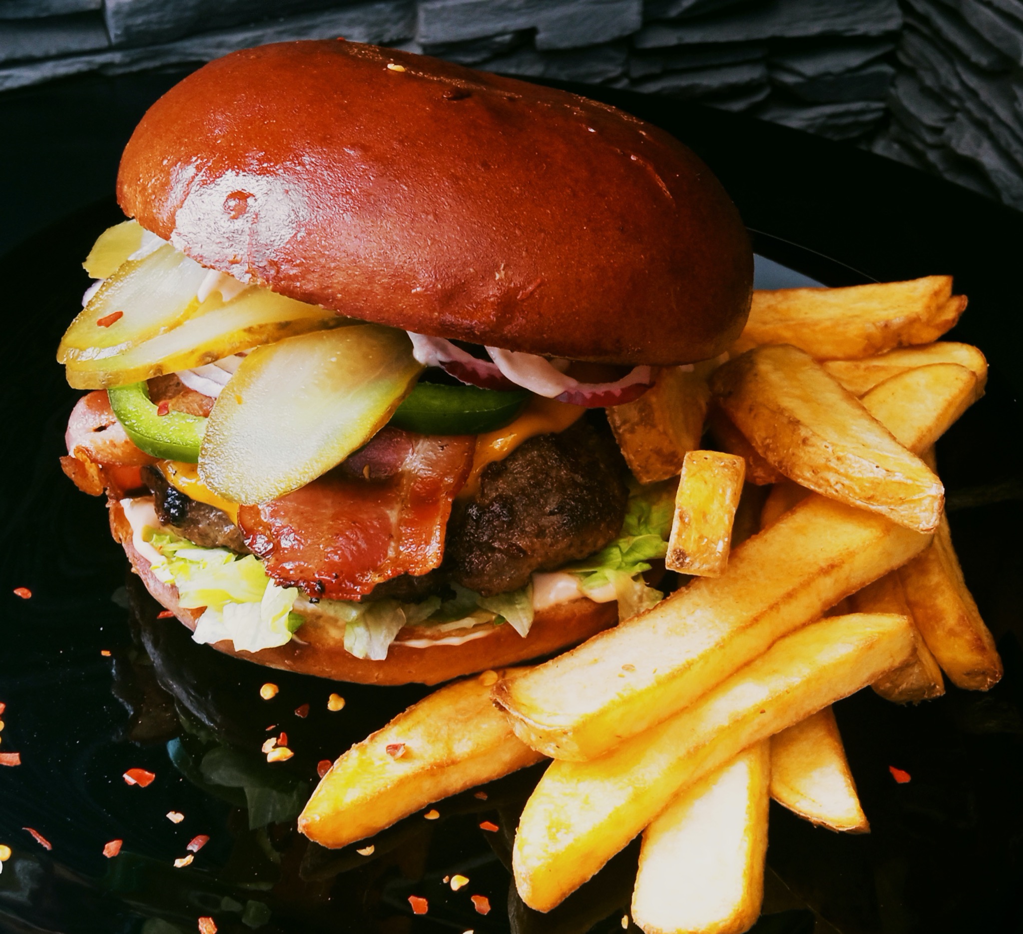 Soczysty hamburger z bekonem, piklami, cebulą i jalapeño, podany z frytkami na czarnym talerzu z posypką z papryczki chili.