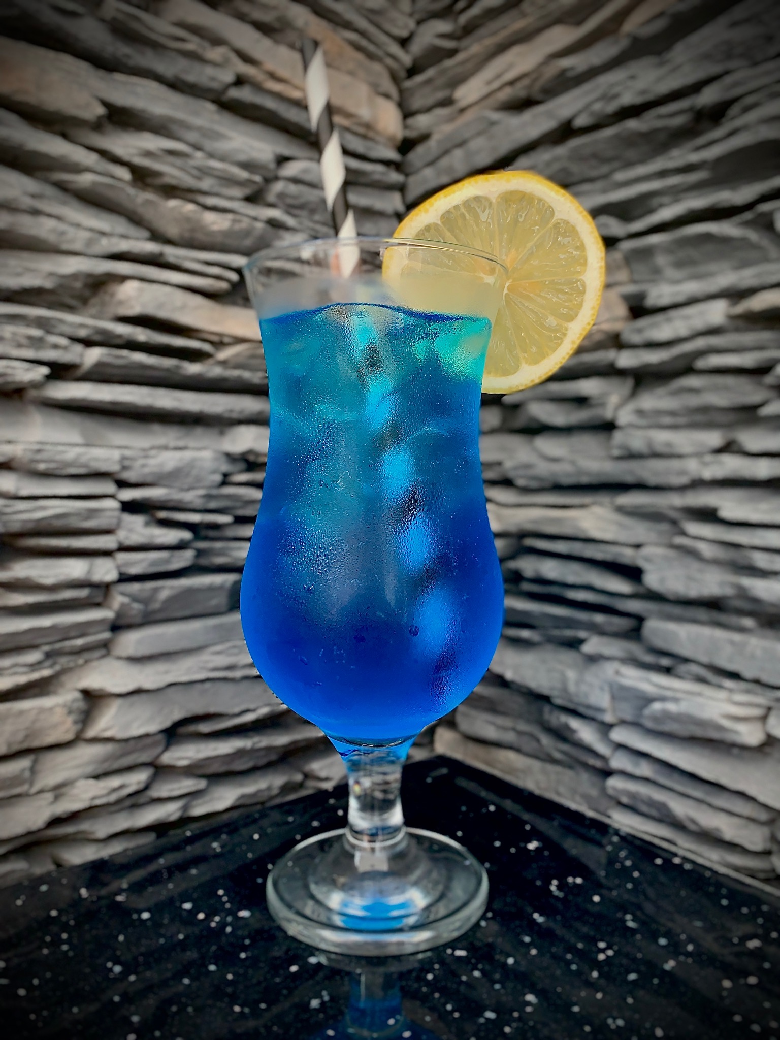 Orzeźwiający, błękitny drink z lodem, słomką w czarno-białe paski i plasterkiem cytryny na tle kamiennej ściany.