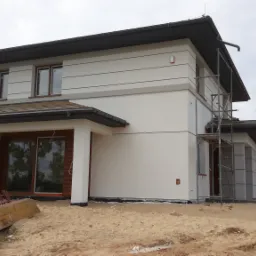 Dwukondygnacyjny dom z jasną elewacją w trakcie wykończenia, widoczne rusztowania i materiały budowlane na piaszczystym terenie.