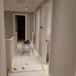 Korytarz w trakcie odświeżania, zabezpieczona podłoga taśmą malarską, drabina rozstawiona na środku, ściany pomalowane na szaro, widoczne otwarte drzwi do pomieszczeń.