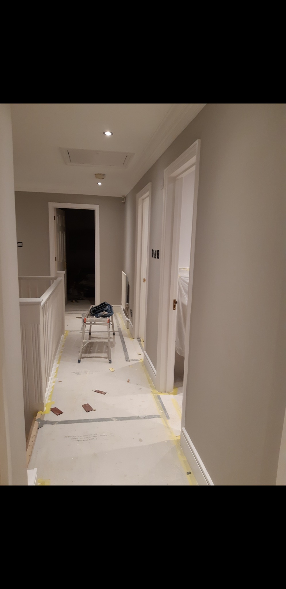 Korytarz w trakcie odświeżania, zabezpieczona podłoga taśmą malarską, drabina rozstawiona na środku, ściany pomalowane na szaro, widoczne otwarte drzwi do pomieszczeń.