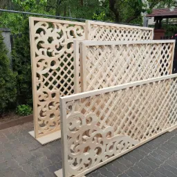 Surowe, drewniane panele z kratką i dekoracyjnymi wycięciami, ustawione na kostce brukowej w ogrodzie, czekające na montaż.