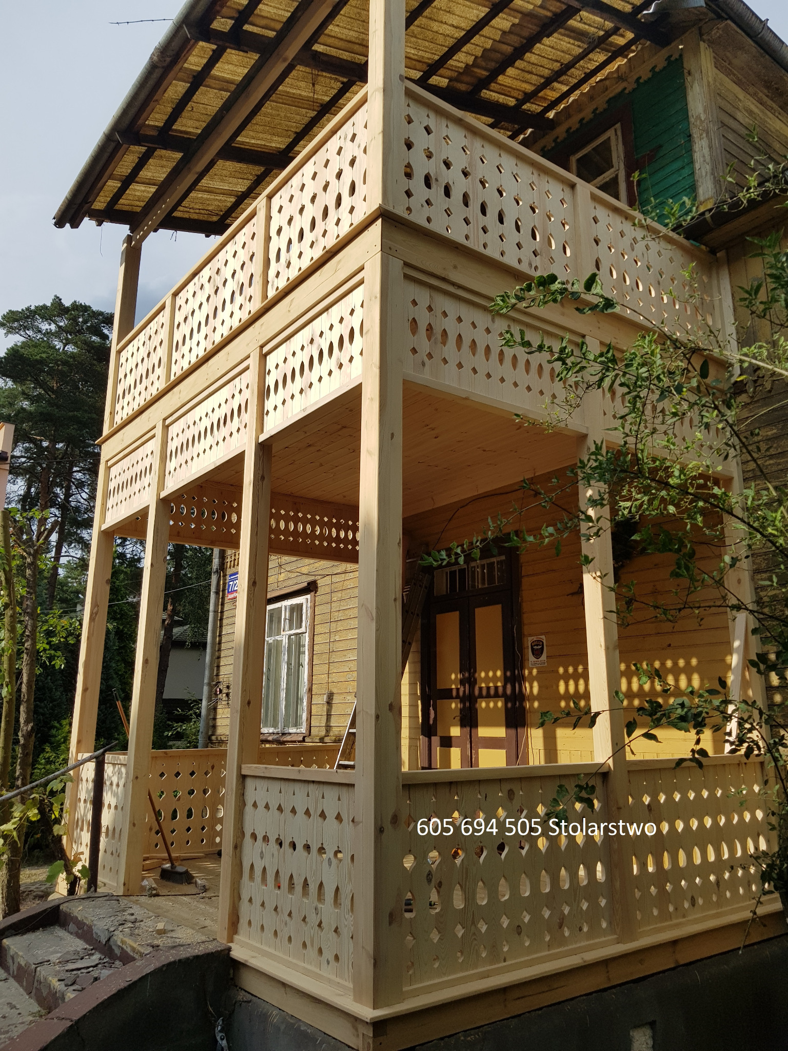 Nowa, dwupoziomowa weranda z surowego drewna z dekoracyjnymi wycięciami, przylegająca do starego, drewnianego domu z żółtą elewacją. Widoczny numer telefonu i napis 'Stolarstwo' na dolnej...