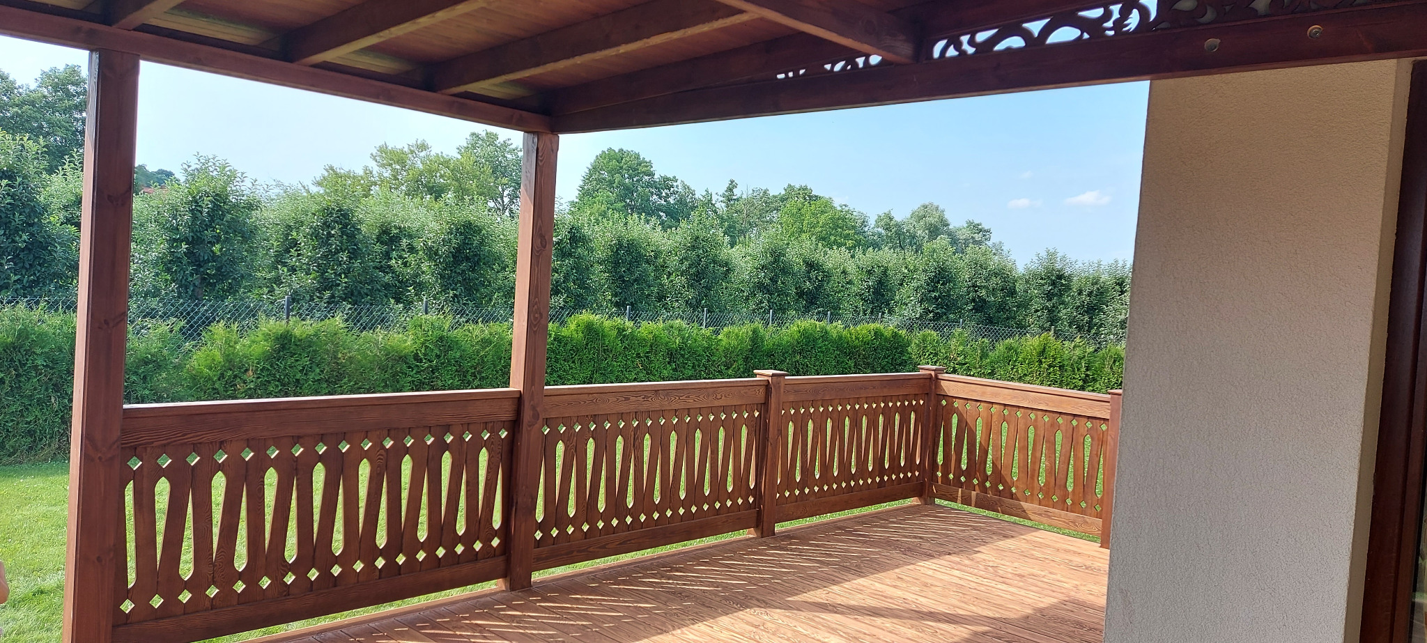Zadaszony taras z drewnianą podłogą i ozdobną balustradą, widok na zielony ogród z drzewami i krzewami.