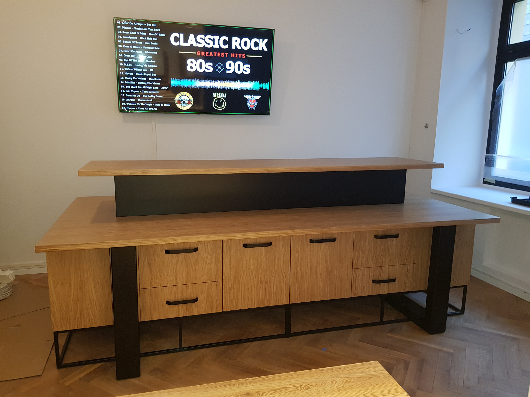 Drewniana komoda z czarnymi, metalowymi nogami i uchwytami, stojąca na parkiecie w jodełkę, z telewizorem wyświetlającym listę utworów rockowych w tle.