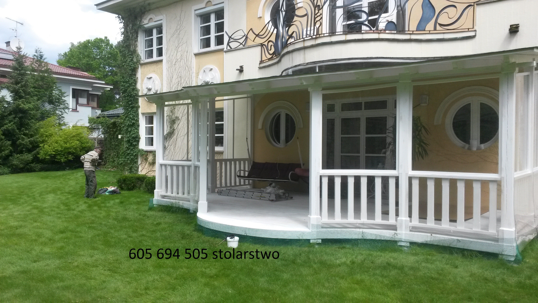 Biała, częściowo przeszklona konstrukcja tarasowa przylegająca do budynku z charakterystycznymi okrągłymi oknami, widoczny fragment trawnika i pracownik w tle, na trawie widoczny numer telefonu...