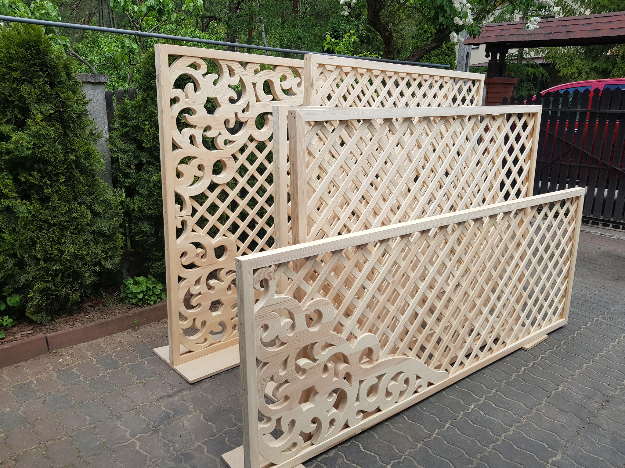 Surowe, drewniane panele z kratką i dekoracyjnymi wycięciami, ustawione na kostce brukowej w ogrodzie, czekające na montaż.