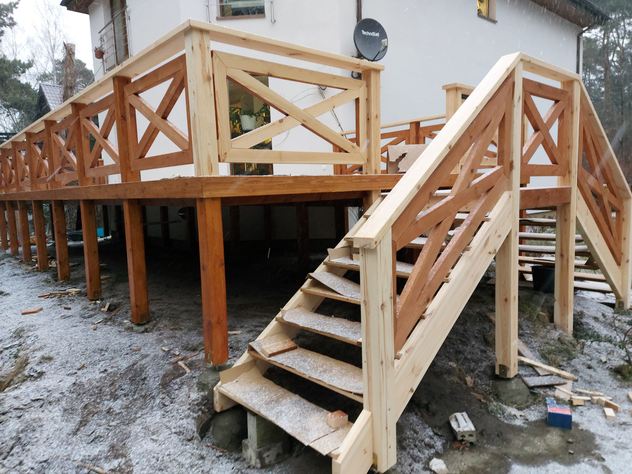 Konstrukcja drewnianego tarasu z balustradą w trakcie budowy, widoczne schody i elementy mocujące, lekko ośnieżone otoczenie.