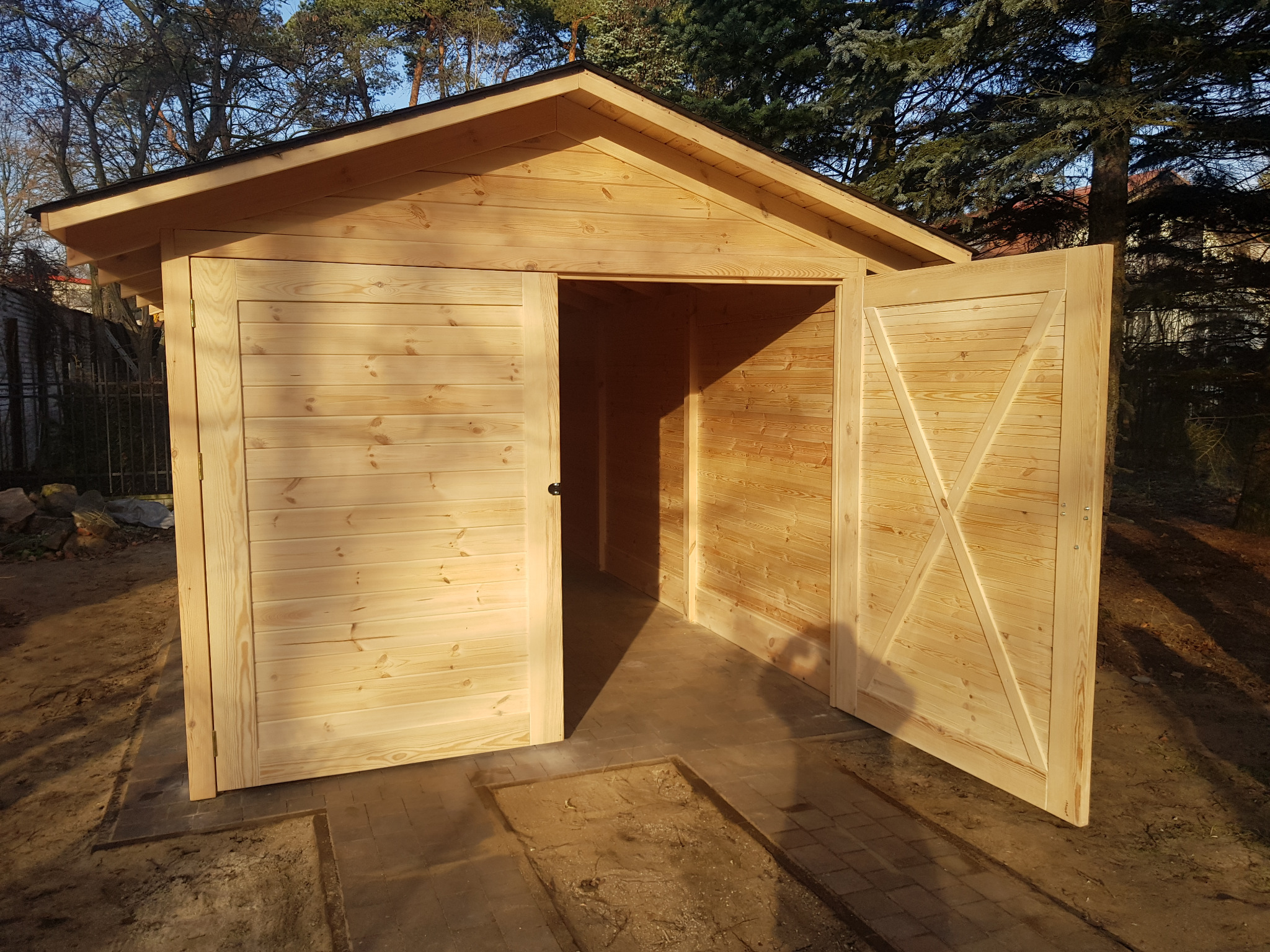 Nowa, surowa drewniana altana z otwartymi dwuskrzydłowymi drzwiami, widoczne ślady po niedawnej instalacji w postaci wykopanych dołów w ziemi.