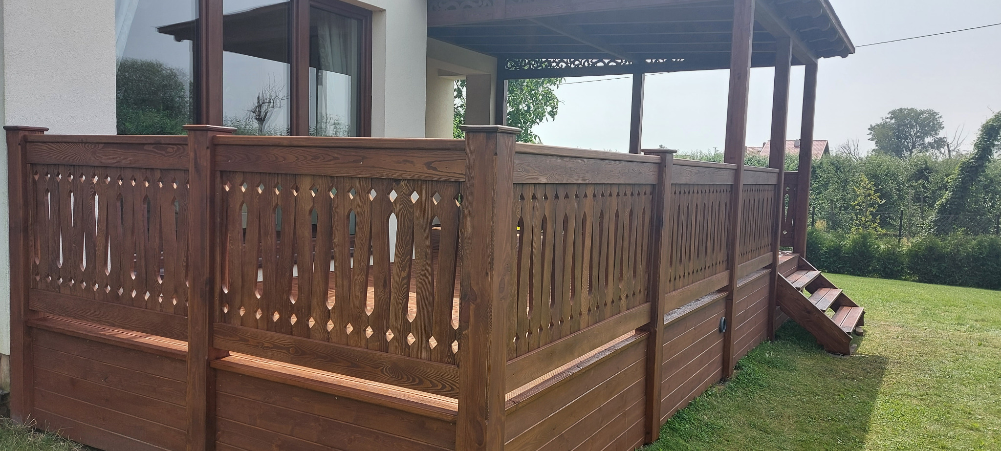 Zadaszony taras z drewnianą balustradą o dekoracyjnym wzorze, schody wejściowe na taras, widok na ogród.