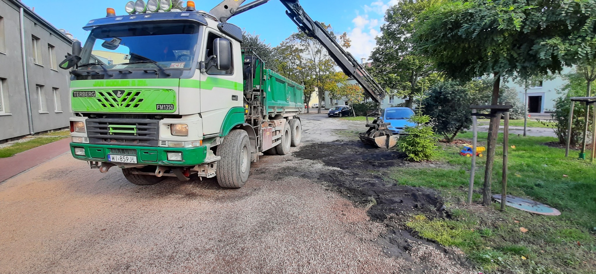 Ciężarówka z dźwigiem Terberg FM1350 z zielono-białym malowaniem, nabierająca ziemię na trawniku w otoczeniu drzew i budynków.