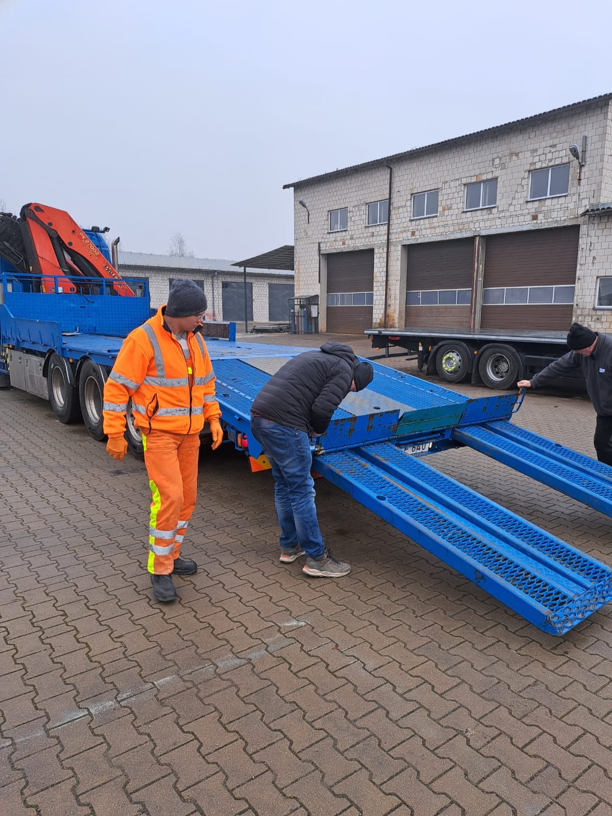 Dwóch mężczyzn przygotowuje niebieską lawetę z pomarańczowym dźwigiem do załadunku, jeden w odblaskowym stroju, drugi w kurtce, na tle budynku z bramami garażowymi.