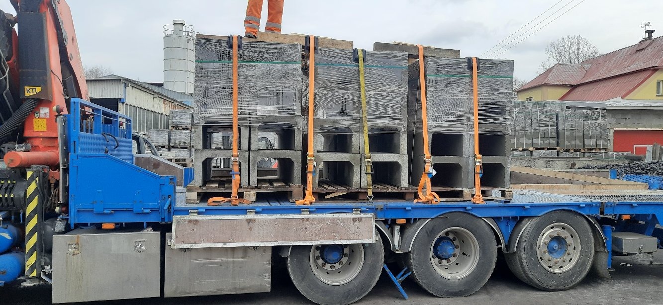 Załadunek betonowych elementów prefabrykowanych o przekroju U na niebieską platformę transportową z pomarańczowymi pasami zabezpieczającymi, widok z boku w pochmurny dzień.