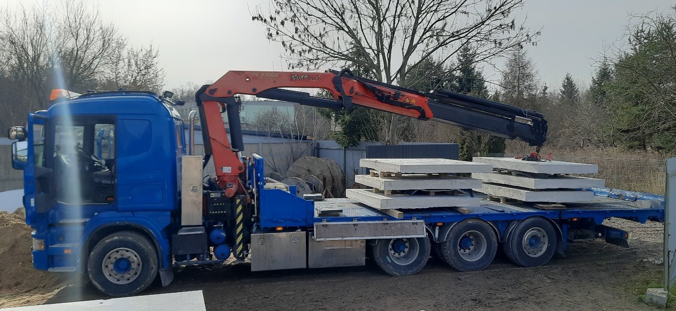 Niebieska ciężarówka z pomarańczowym dźwigiem PALFINGER PK 27002 przewozi betonowe płyty na paletach, widoczne drzewa w tle.