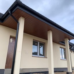 ArDrew - Beżowy dom jednorodzinny z brązową podbitką dachową i czarnymi rynnami, widok na fragment elewacji z oknami i kolumnami podtrzymującymi zadaszenie, teren wokół domu w trakcie prac wykończeniowych.