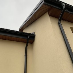 ArDrew - Fragment elewacji domu z zamontowanym systemem rynnowym w kolorze czarnym, widoczne drewniane podbitki dachowe.