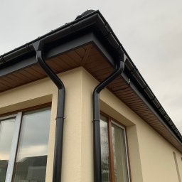 ArDrew - Fragment elewacji budynku z zamontowanym systemem rynnowym w kolorze czarnym, widoczne brązowe podbitki dachowe, kremowa elewacja i okna z brązowymi ramami.