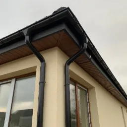 Fragment elewacji budynku z zamontowanym systemem rynnowym w kolorze czarnym, widoczne brązowe podbitki dachowe, kremowa elewacja i okna z brązowymi ramami.