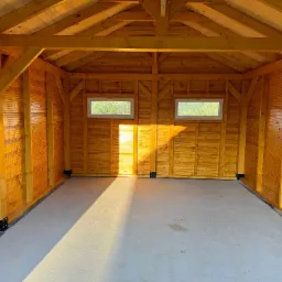 Wnętrze nowej, drewnianej konstrukcji z dwoma oknami, betonową posadzką i widocznymi elementami więźby dachowej, oświetlone wpadającym światłem słonecznym.