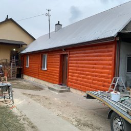 ArDrew - Dom z nową, pomarańczową elewacją drewnianą, widok z zewnątrz podczas prac wykończeniowych; w tle widoczny drugi budynek, na pierwszym planie narzędzia i przyczepa.