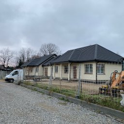 ArDrew - Budowa szeregu domów jednorodzinnych z ciemną dachówką, widoczny biały bus i pomarańczowa koparka za ogrodzeniem z siatki, teren wysypany żwirem, pochmurne niebo.