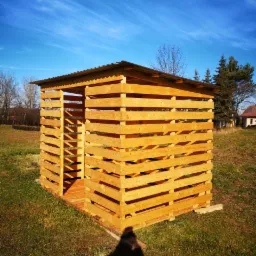 Konstrukcja z jasnego drewna, prawdopodobnie drewutnia, z metalowym dachem, widziana w słoneczny dzień na trawiastym terenie z drzewami w tle. Na pierwszym planie cień osoby robiącej zdjęcie.