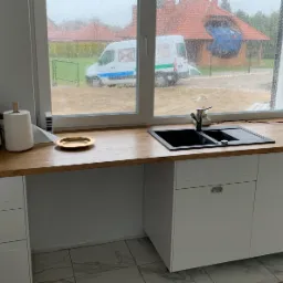 Nowoczesna kuchnia z drewnianym blatem, białymi szafkami z metalowymi uchwytami i czarnym zlewozmywakiem, widok na zewnątrz na zewnątrz przez okno w deszczowy dzień, gdzie widać zaparkowany biały bus.