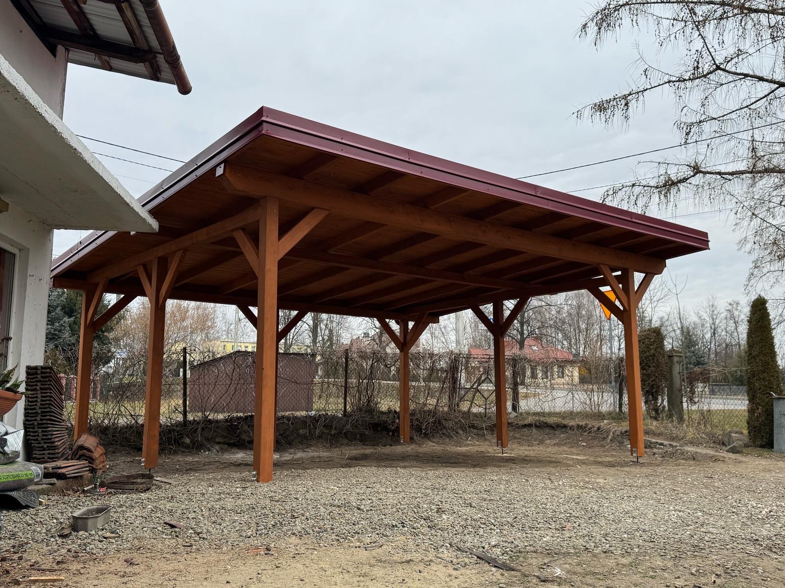 Zadaszenie z drewna o czerwonym dachu na żwirowym podłożu, widoczne elementy konstrukcyjne i dom w tle.
