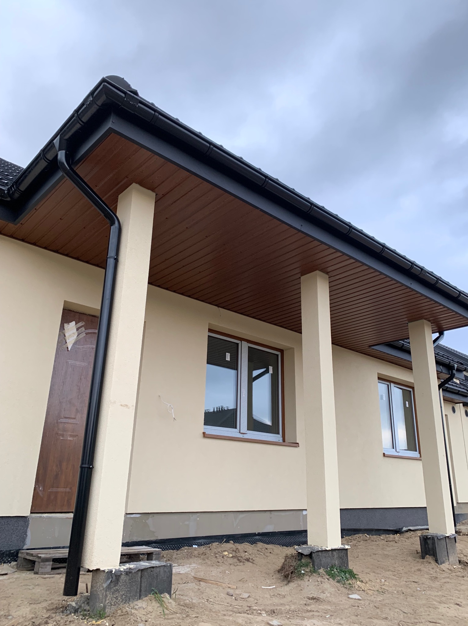 Beżowy dom jednorodzinny z brązową podbitką dachową i czarnymi rynnami, widok na fragment elewacji z oknami i kolumnami podtrzymującymi zadaszenie, teren wokół domu w trakcie prac wykończeniowych.