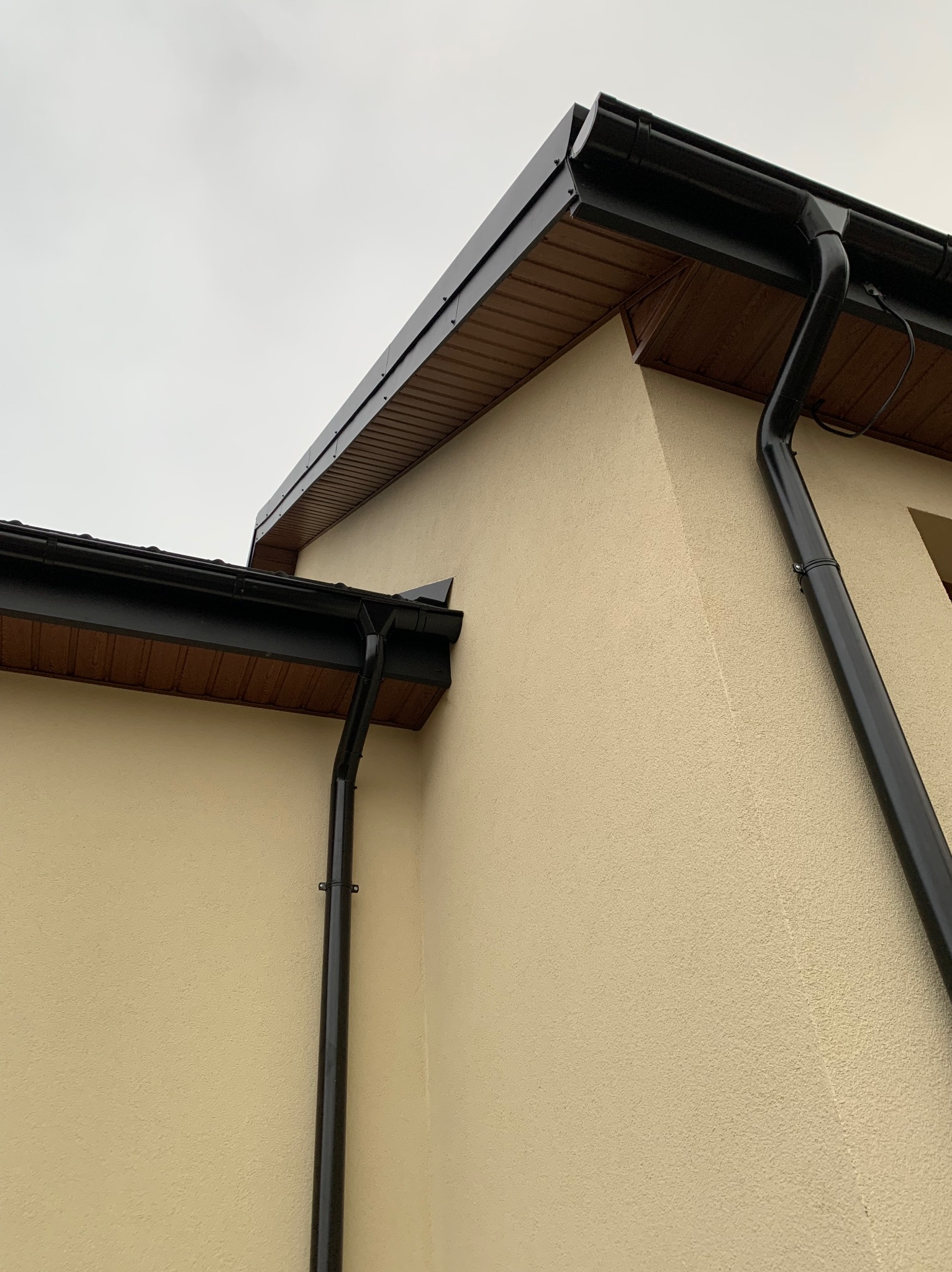 Fragment elewacji domu z zamontowanym systemem rynnowym w kolorze czarnym, widoczne drewniane podbitki dachowe.