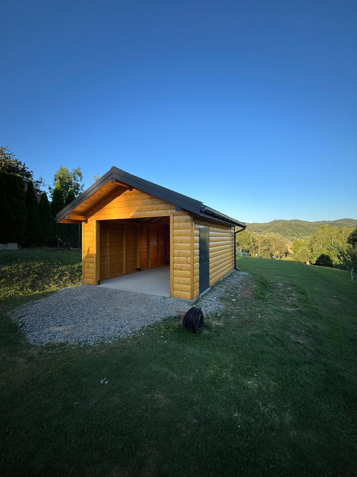 Drewniana konstrukcja garażowa z płaskim dachem na żwirowym podłożu, z czarną rurą zwiniętą obok, na tle zielonego wzgórza i błękitnego nieba.