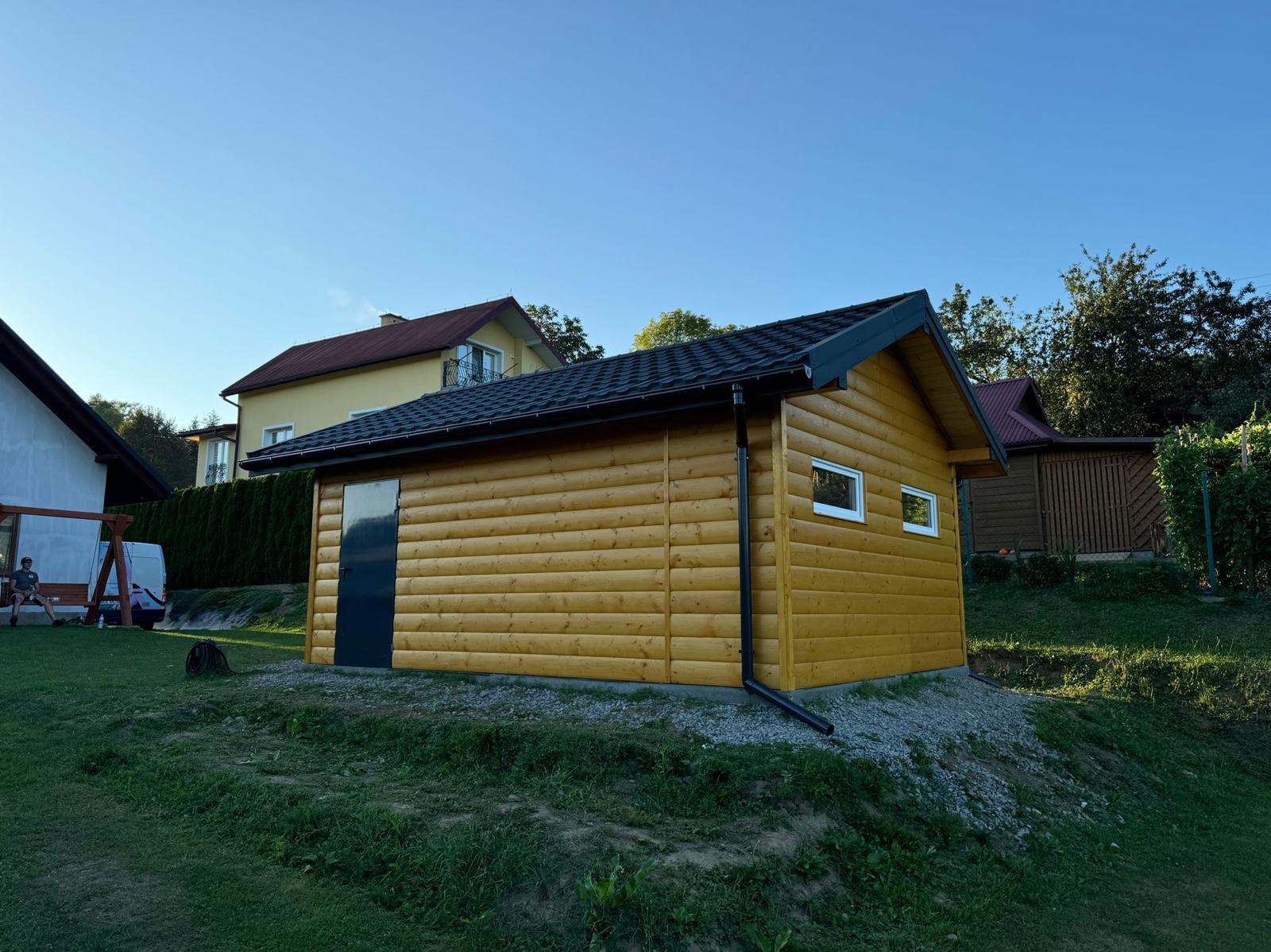 Żółta, nowa konstrukcja z drewna, z ciemną, metalową dachówką i czarną rynną, na nierównym terenie z kamieniami i trawą, w tle domy i drzewa.